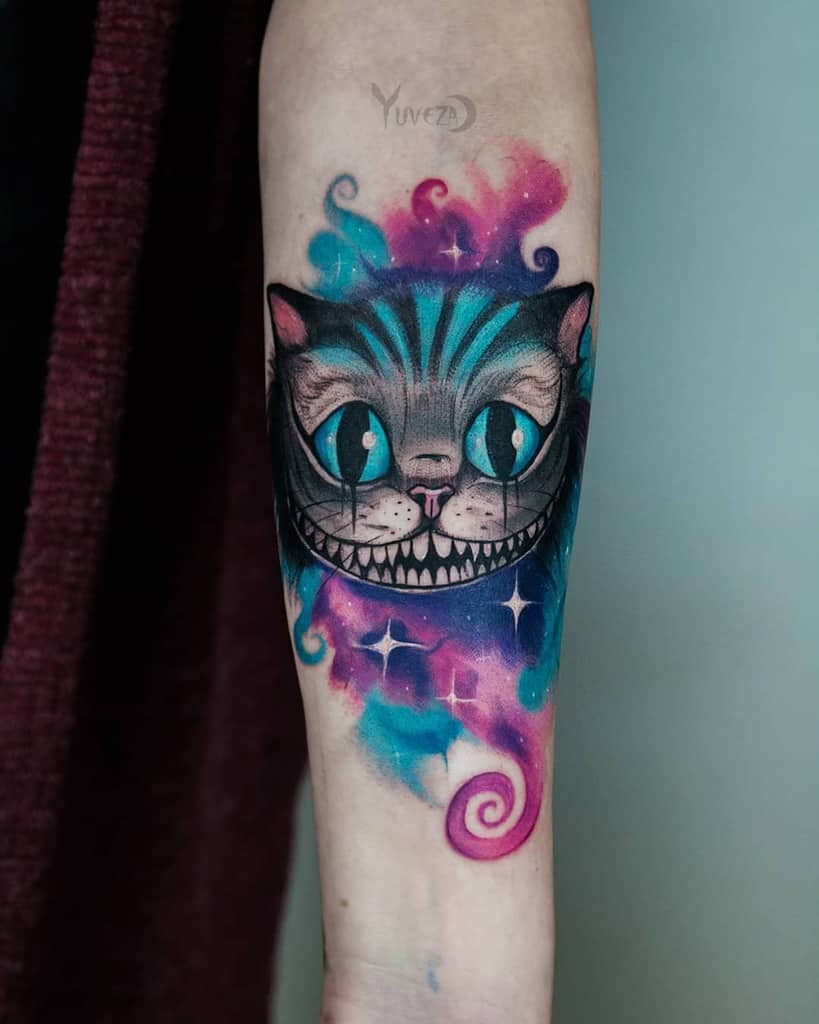 Tattoo of a grinning Cheshire cat with colorful, swirling background on the inner forearm