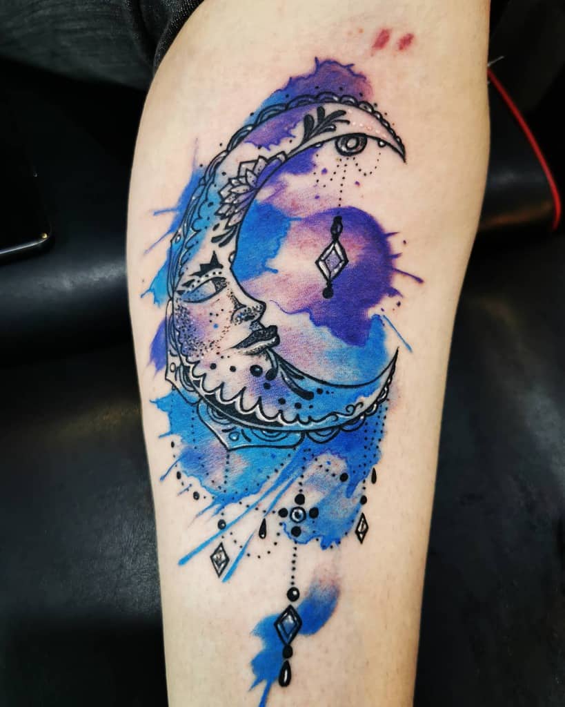 Tattoo of a crescent moon adorned with watercolor blue and purple splashes on an arm