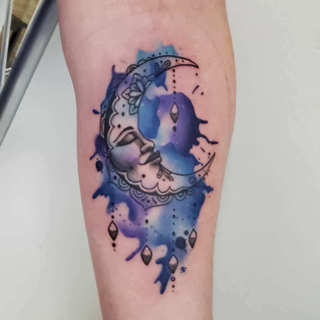 Tattoo of a crescent moon with a face amidst a blue and purple watercolor background, adorned with delicate hanging gems