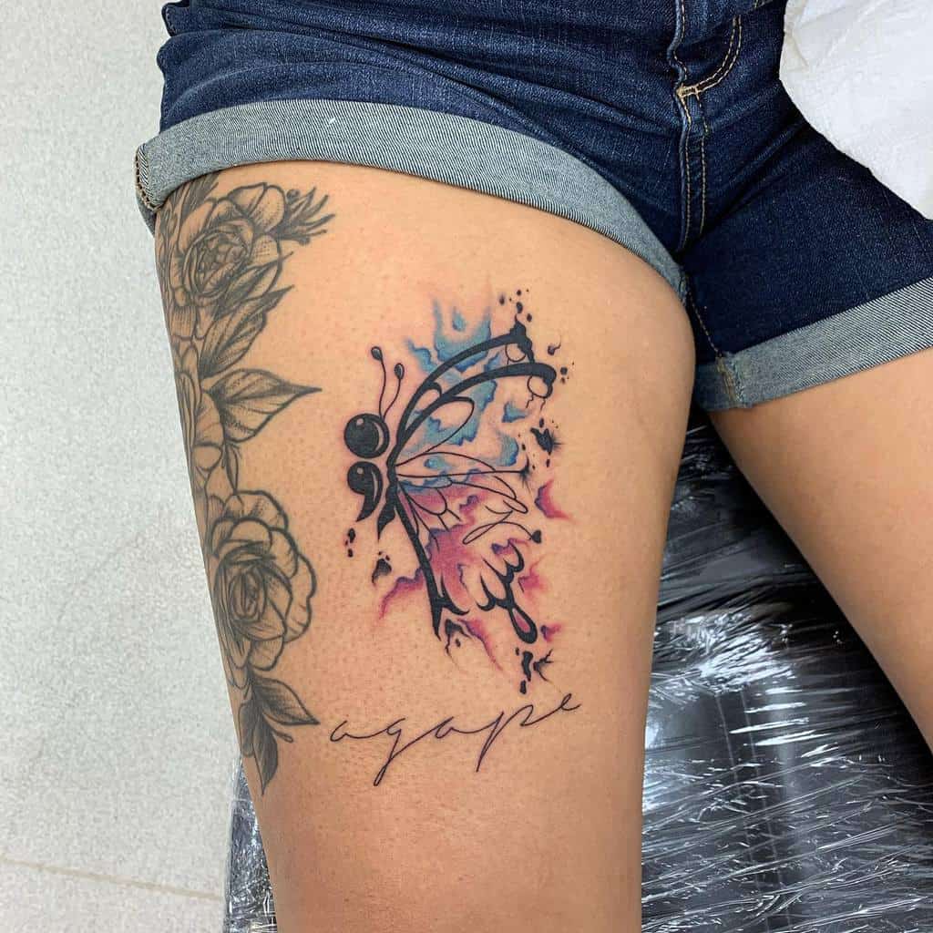 A watercolor semicolon butterfly tattoo on the thigh, with the word 'agape' below, symbolizing unconditional love and mental health awareness