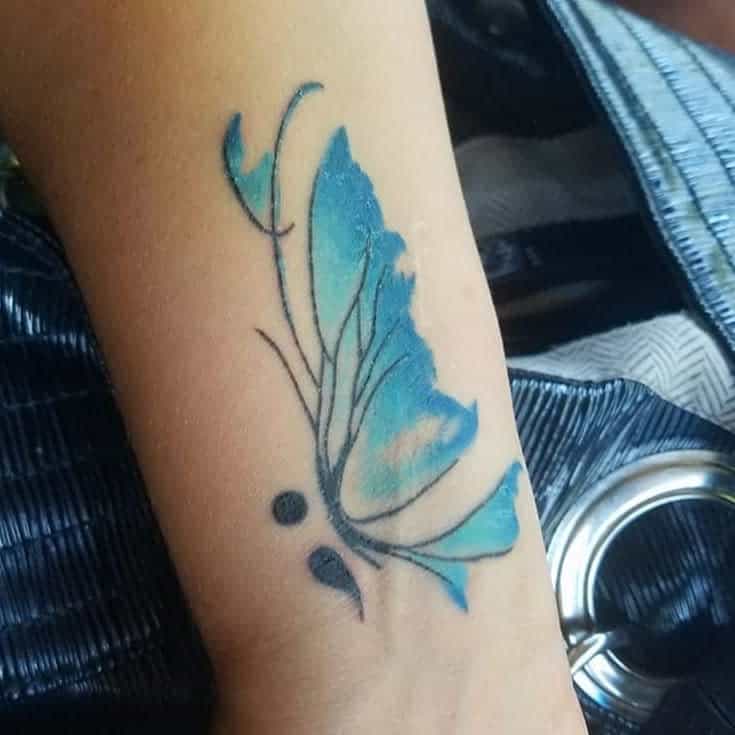 A delicate blue watercolor semicolon butterfly tattoo on the forearm, symbolizing mental health awareness and personal growth