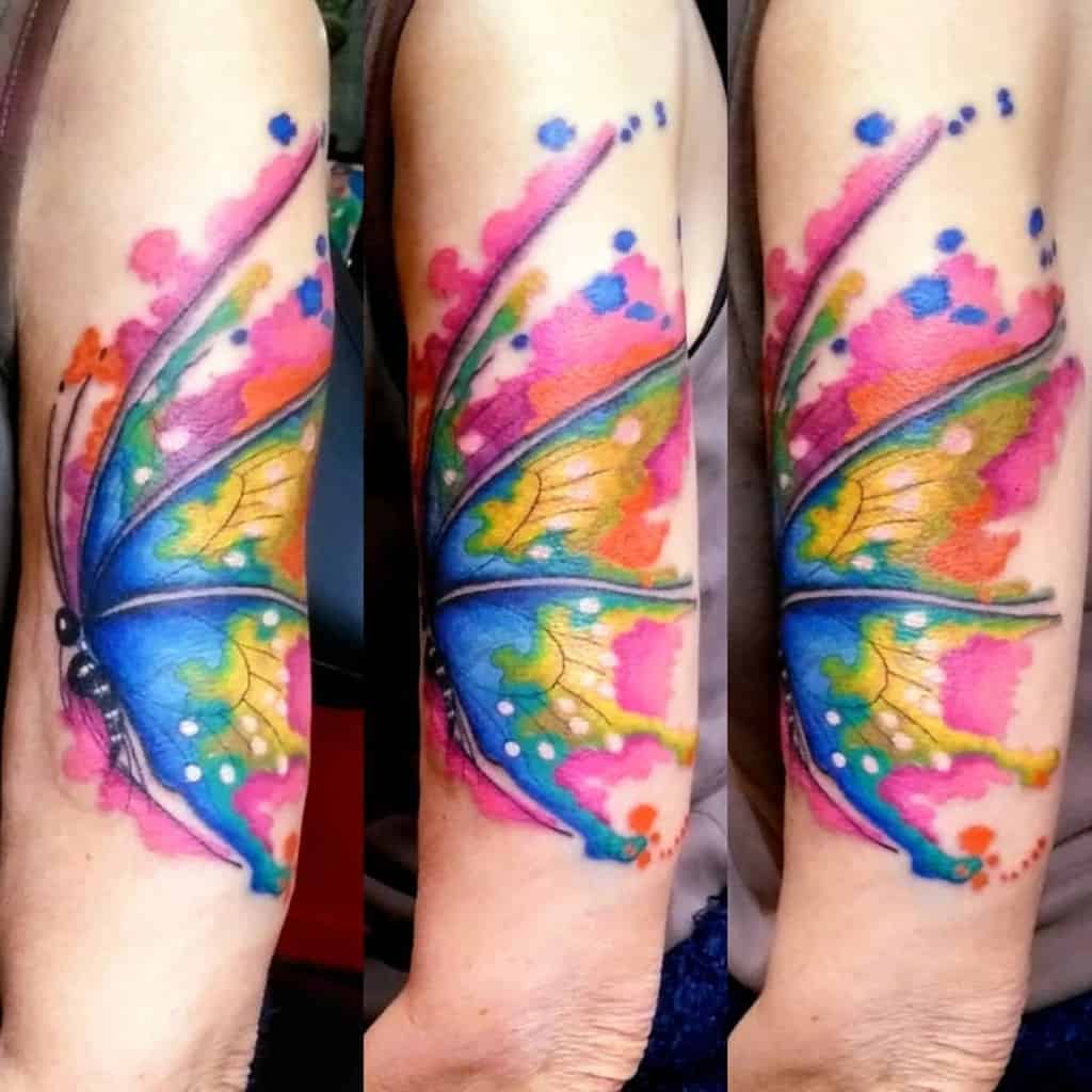 A large, vibrant watercolor butterfly tattoo with a semicolon body, featuring bright splashes of color, symbolizing transformation and mental health awareness