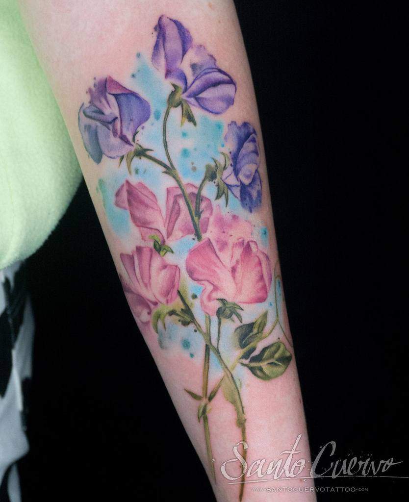 Watercolor-style sweet pea flower tattoo with vibrant pink and purple petals on the forearm. Light blue accents surround the flowers, adding a dreamy, artistic touch