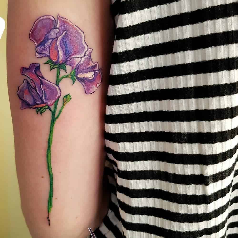 Watercolor sweet pea flower tattoo with bold purple petals and a bright green stem, contrasting beautifully against the skin. The vibrant design adds a modern artistic touch to the upper arm.