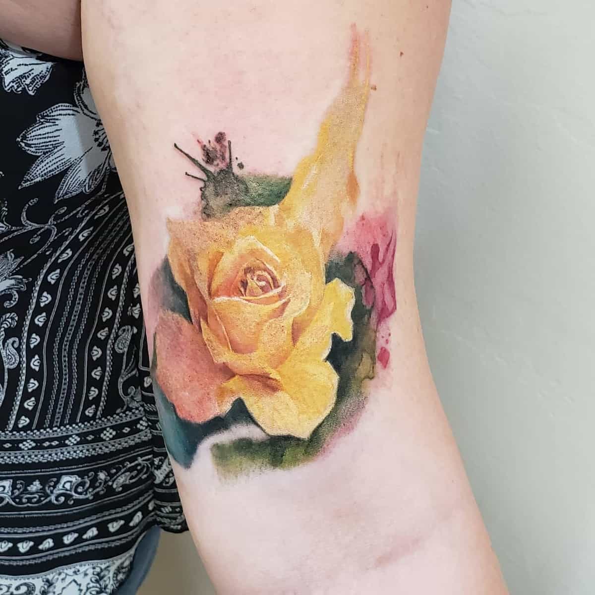 Watercolor-style tattoo of a yellow rose with green leaves and pink accents on the inner arm