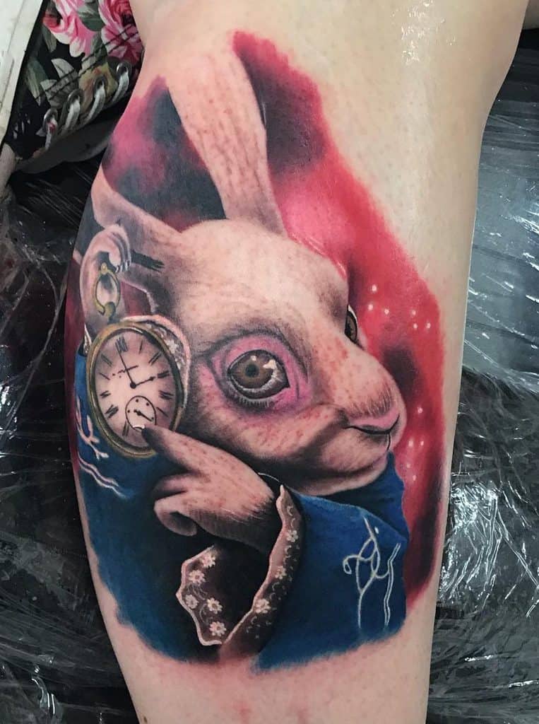 Tattoo of a white rabbit holding a pocket watch, wearing a blue coat with floral sleeves, set against a red background