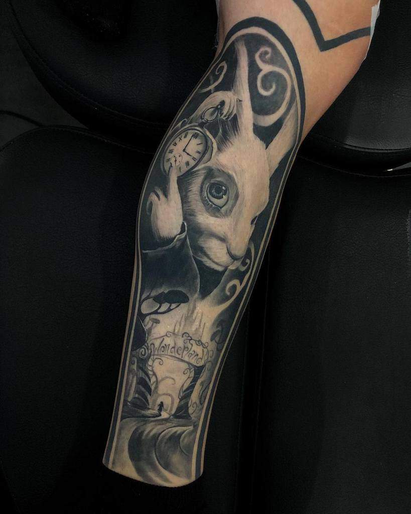 Black and gray tattoo of the White Rabbit from Alice in Wonderland, holding a pocket watch with a detailed background showing mushrooms, swirling smoke, and an archway with 'Wonderland' written on it