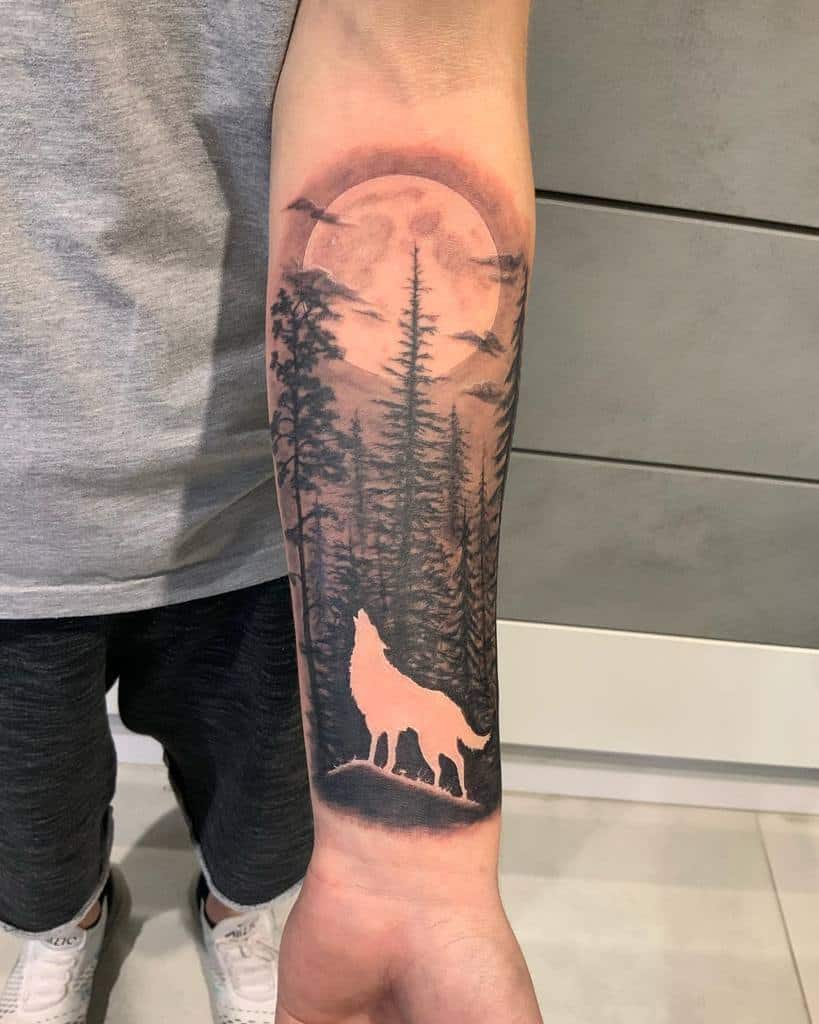 Tattoo of a wolf howling at the moon with trees and birds in the background on a forearm