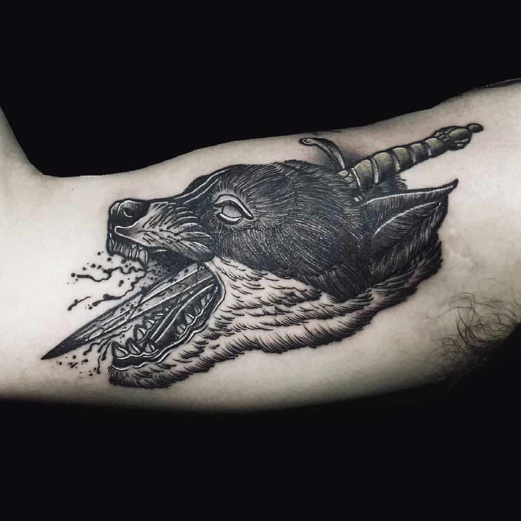 Tattoo of a wolf head with an open mouth, pierced by a dagger, detailed in black and gray ink on a bicep