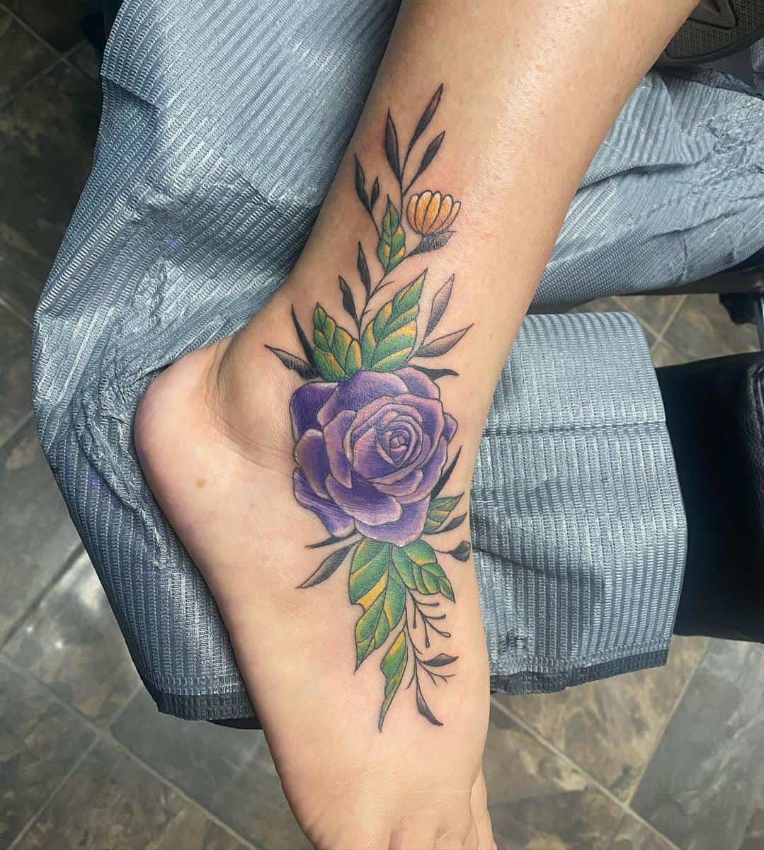 Foot tattoo featuring a large purple rose with green leaves and a small yellow flower, resting on pinstriped fabric