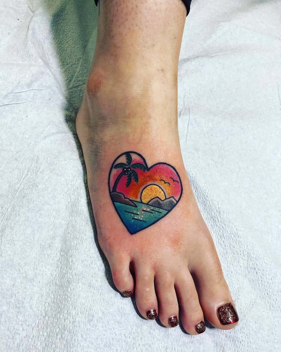 A heart-shaped tattoo with a beach sunset scene on the top of a foot