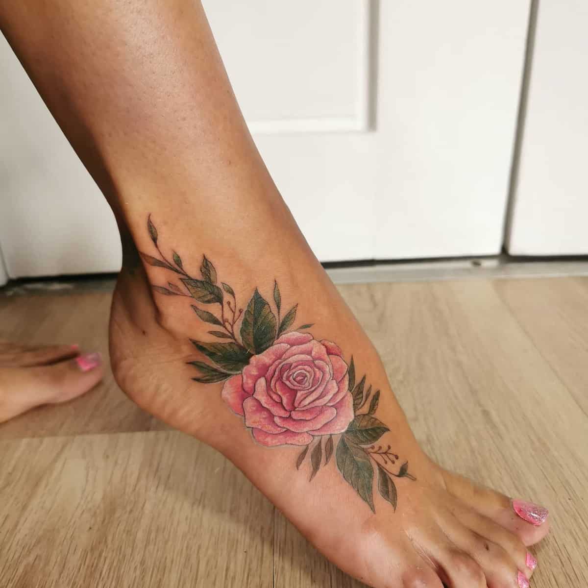 Foot with a rose tattoo on the ankle surrounded by green leaves on a light wood floor