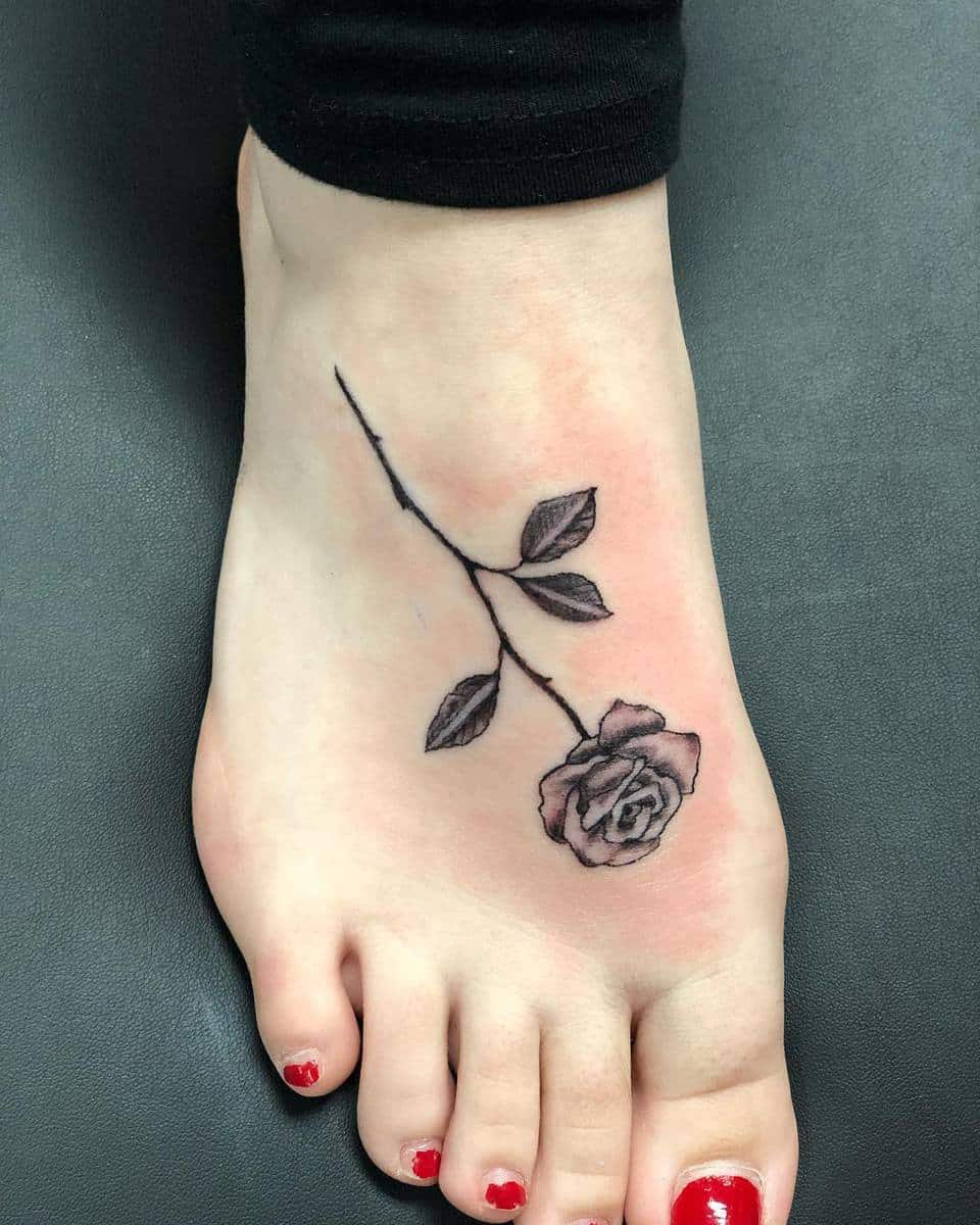 Foot tattoo of a single black rose with a long stem and leaves, located on the top of the foot