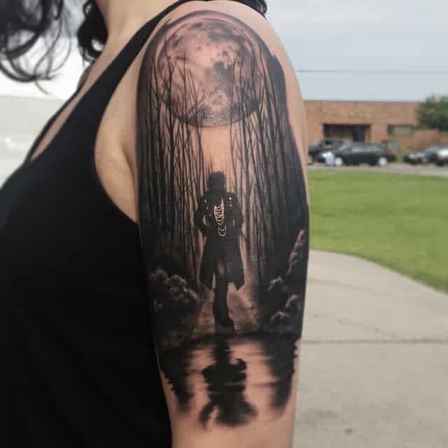 Tattoo of a cloaked figure in woods under a full moon, mirrored in water on upper arm