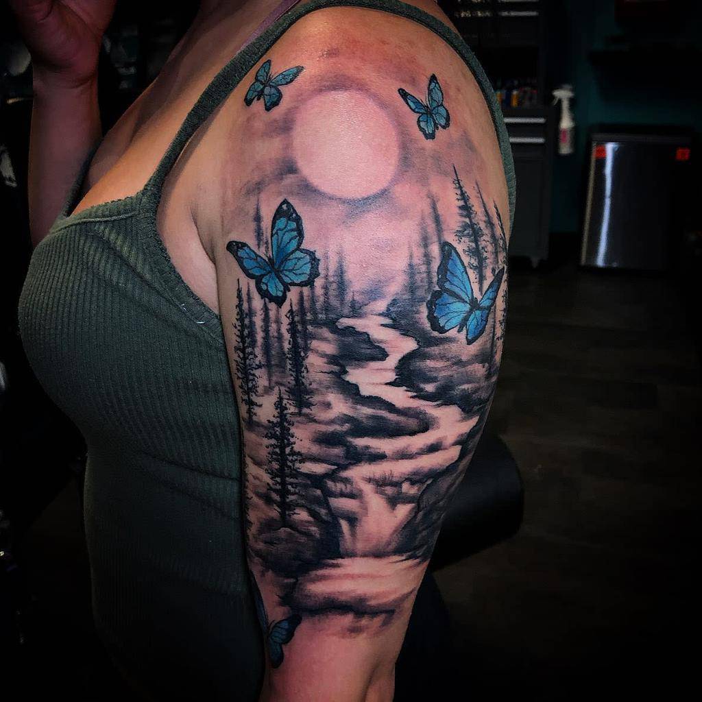 Arm tattoo with a moonlit forest, river, and blue butterflies on the shoulder