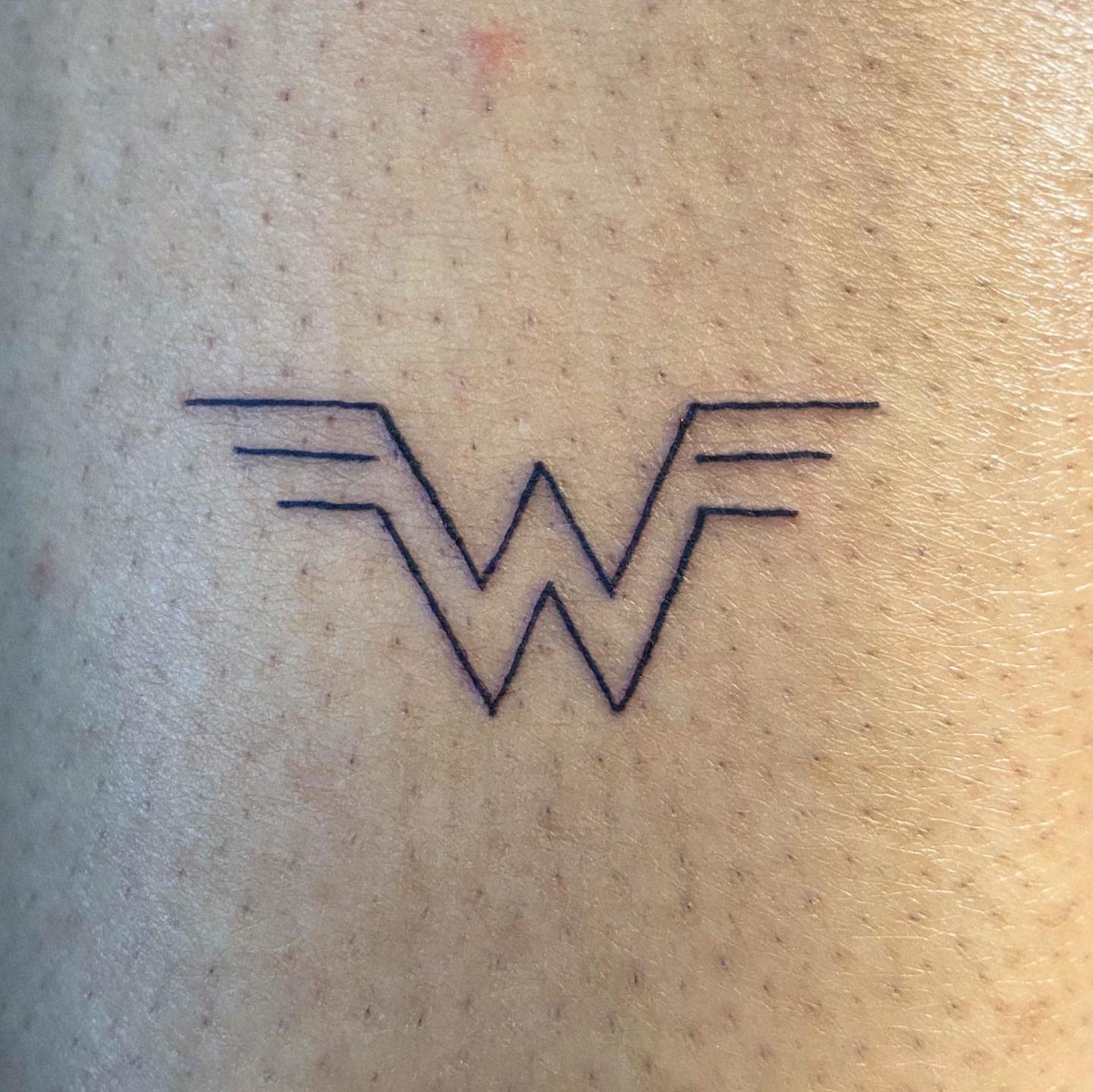 Minimalist tattoo of Wonder Woman's 'W' logo in thin black lines, simple and elegant design