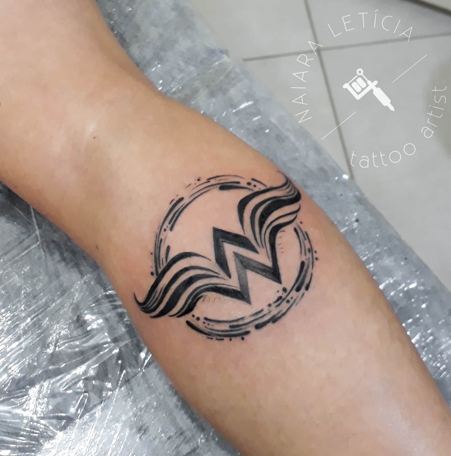 Stylized black ink tattoo of Wonder Woman's 'W' logo with artistic circular accents