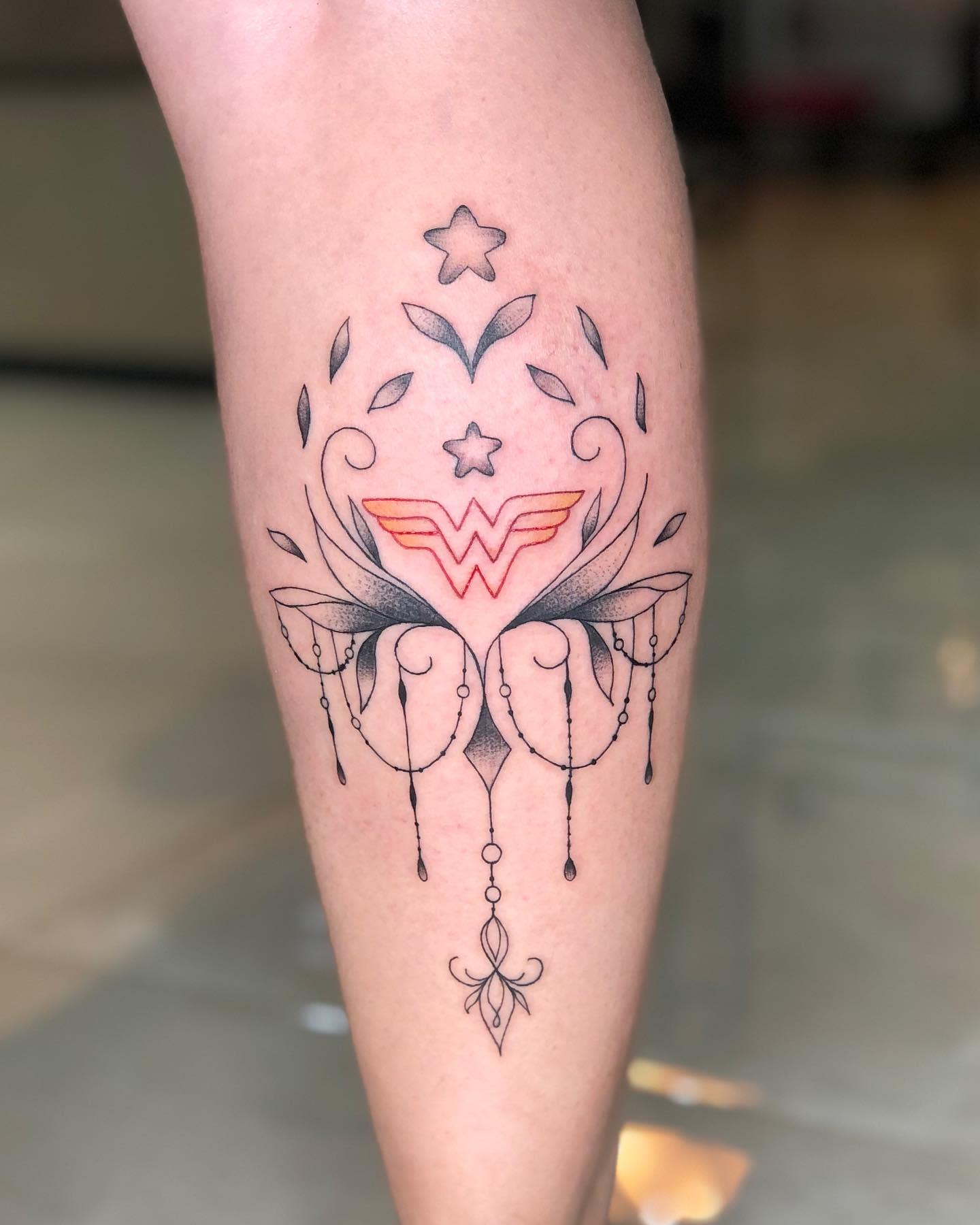 Delicate tattoo of Wonder Woman's 'W' logo with ornate decorative elements and stars