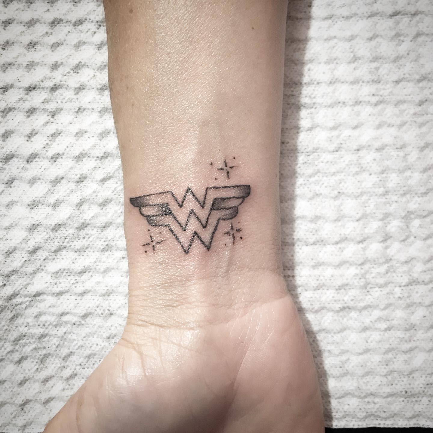 Small Wonder Woman 'W' logo tattoo on wrist, shaded with subtle sparkles surrounding the design