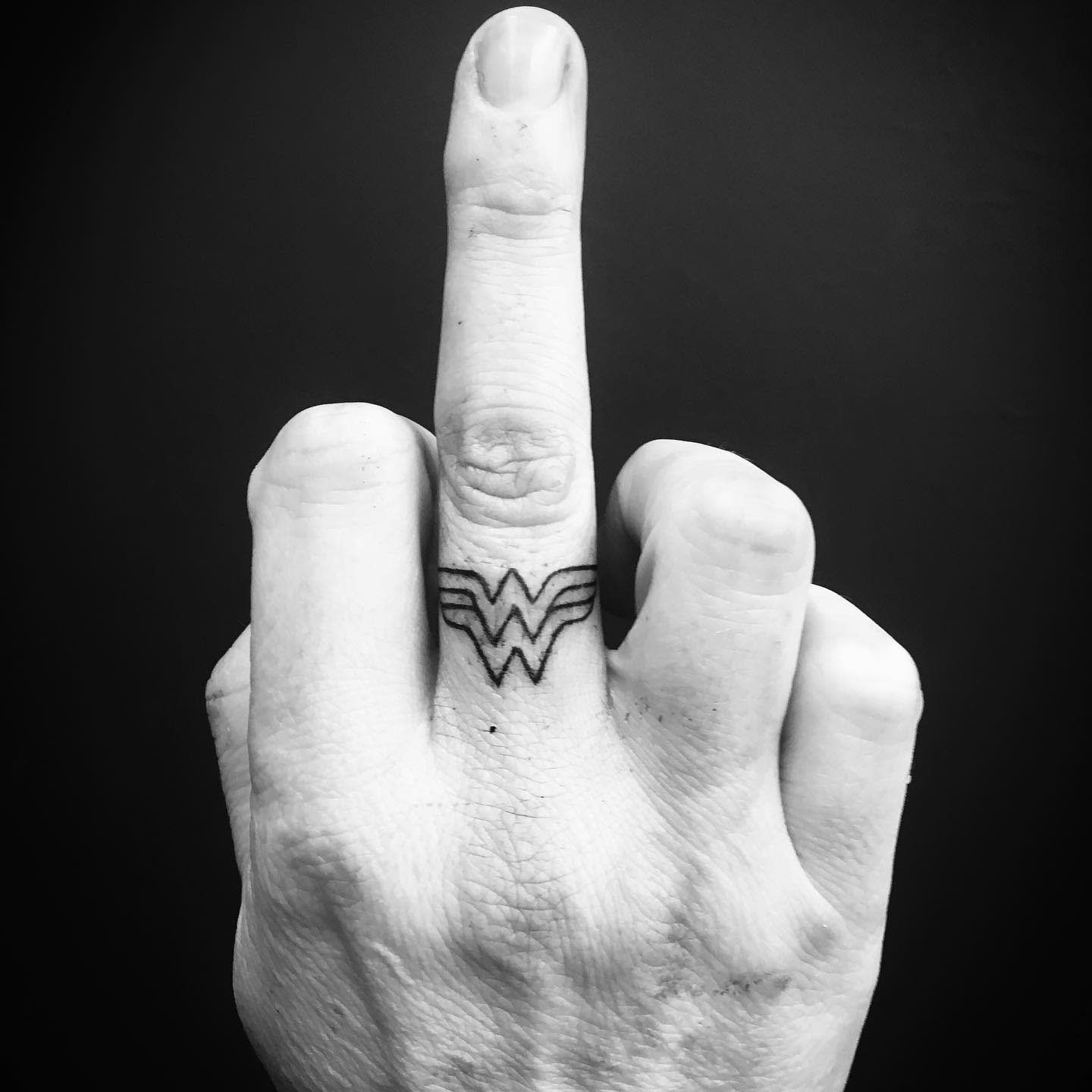Small Wonder Woman 'W' logo tattoo on the middle finger, minimalist design in black ink