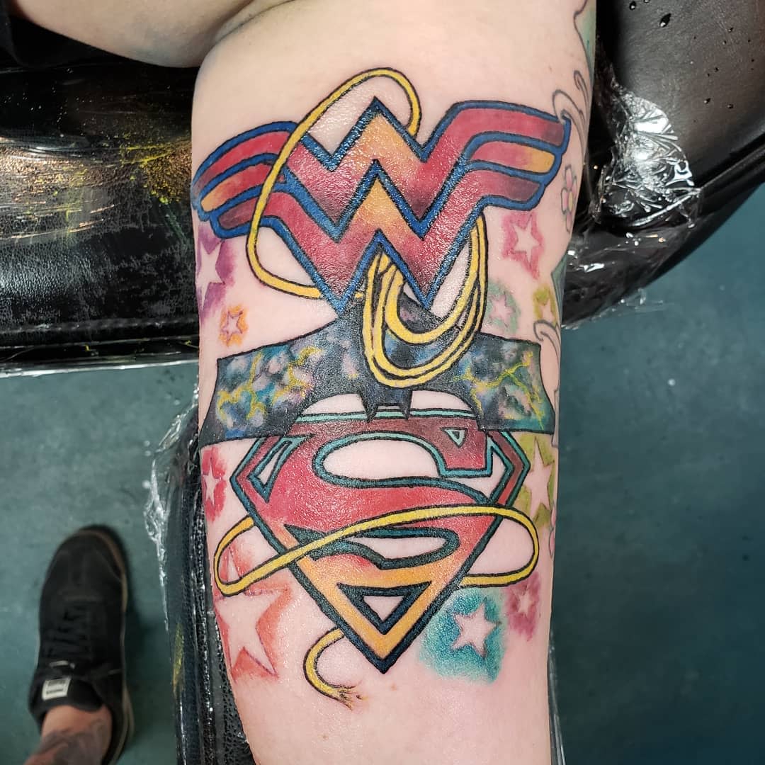 Colorful tattoo combining Wonder Woman's 'W' logo and Superman's 'S' emblem, intertwined with lasso and stars