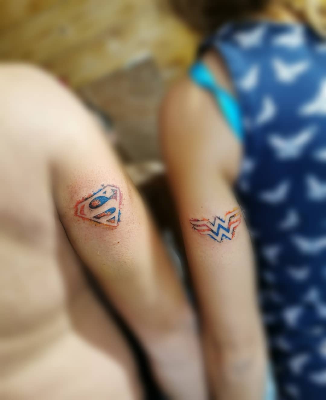 Matching Superman and Wonder Woman logo tattoos on arms, with subtle watercolor effects in red and blue