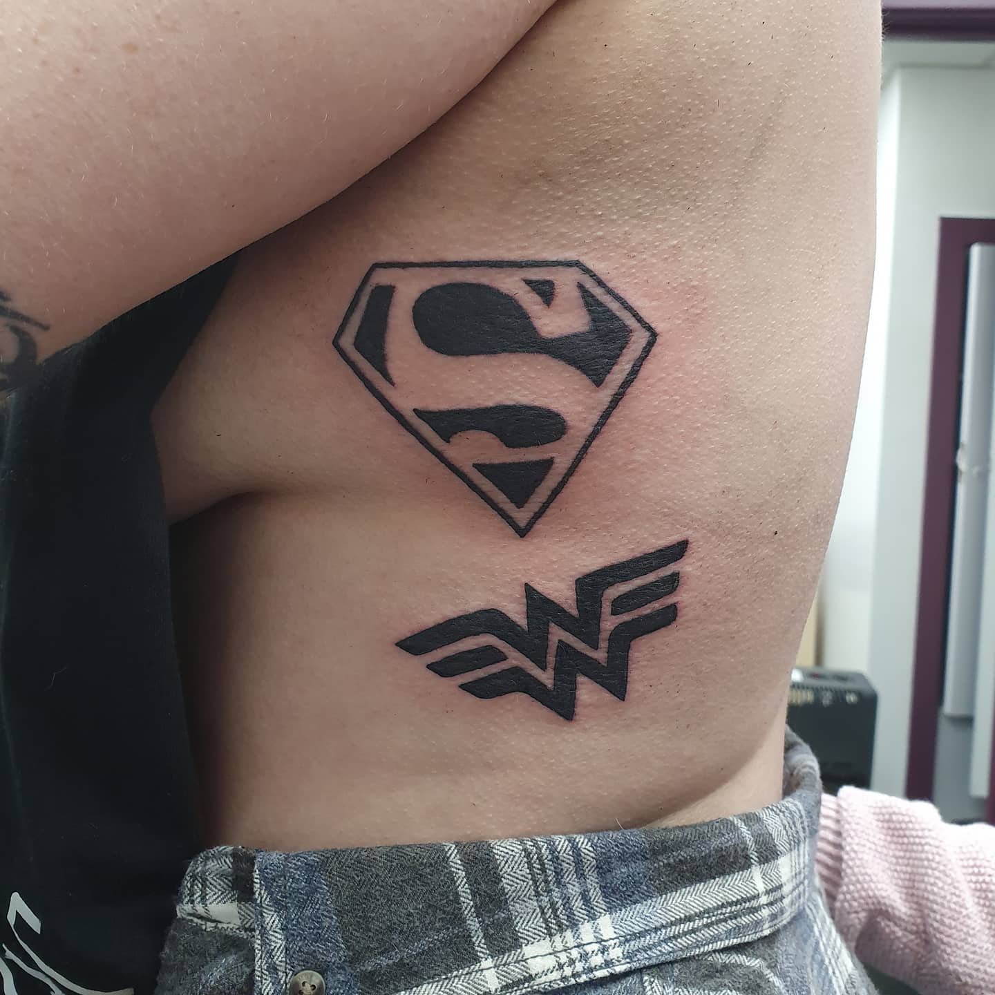 Black ink tattoo featuring Superman's 'S' emblem above Wonder Woman's 'W' logo on the ribcage