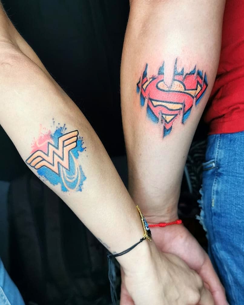 Matching tattoos featuring Superman's 'S' emblem and Wonder Woman's 'W' logo, both with watercolor splashes