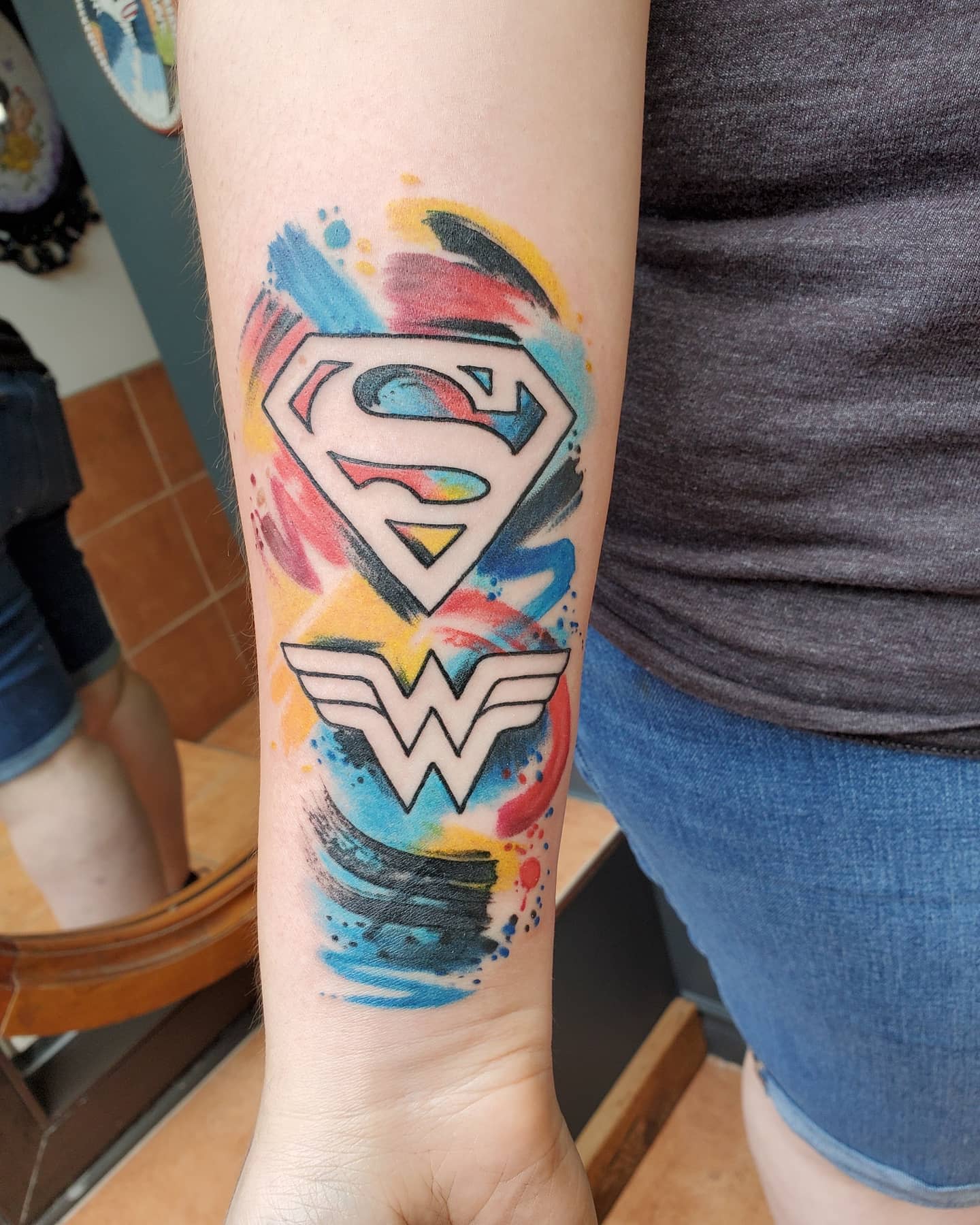 Superman and Wonder Woman logo tattoo with abstract watercolor splashes, combining bold lines and vibrant colors
