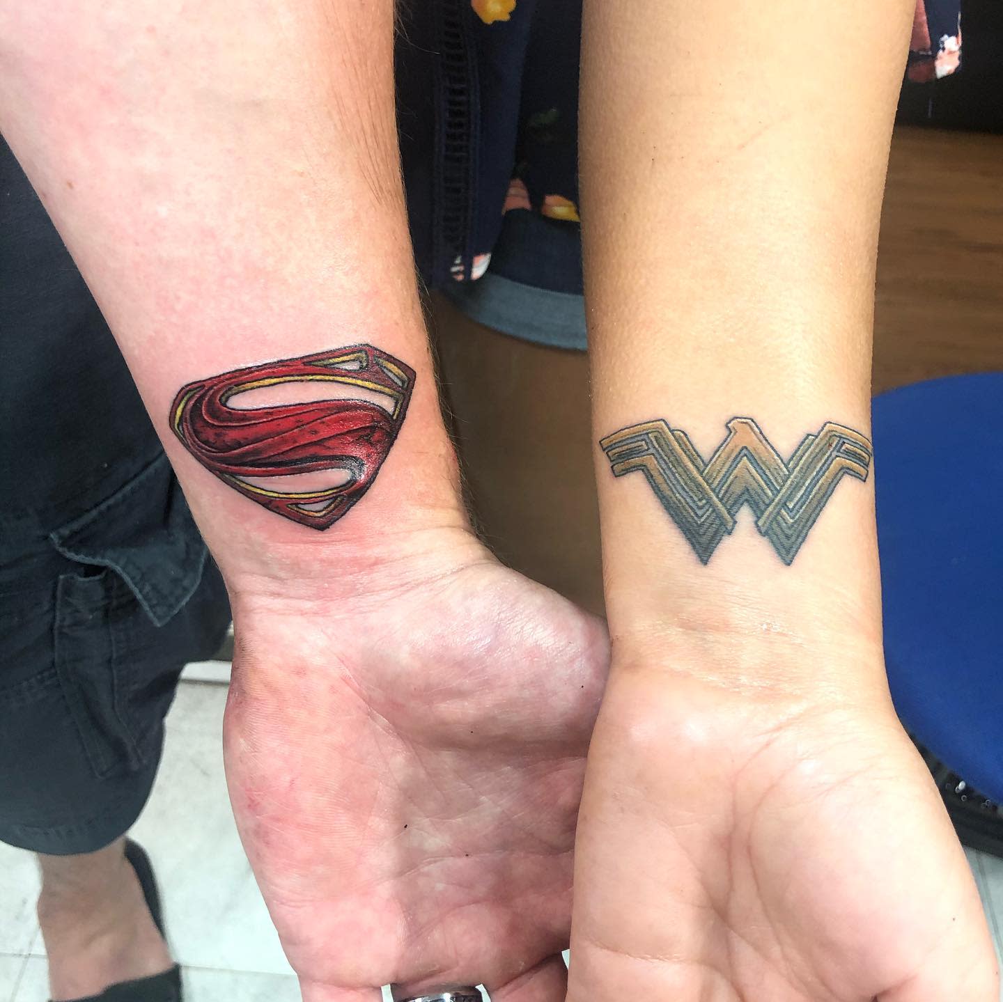 Matching Superman and Wonder Woman wrist tattoos, featuring a detailed red Superman logo and a stylized Wonder Woman logo