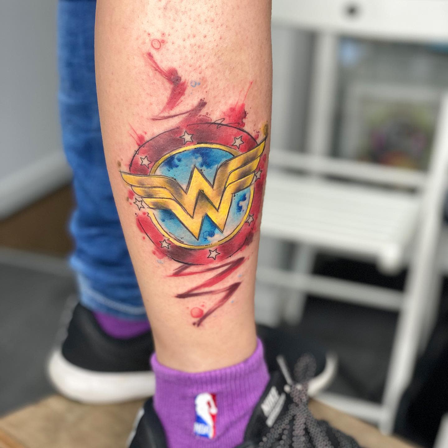Colorful Wonder Woman logo tattoo with a watercolor background, surrounded by stars and dynamic brush strokes