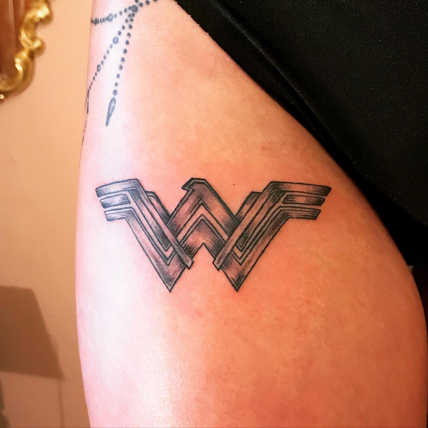 Black and gray Wonder Woman logo tattoo on the thigh, with detailed shading and a bold, clean design