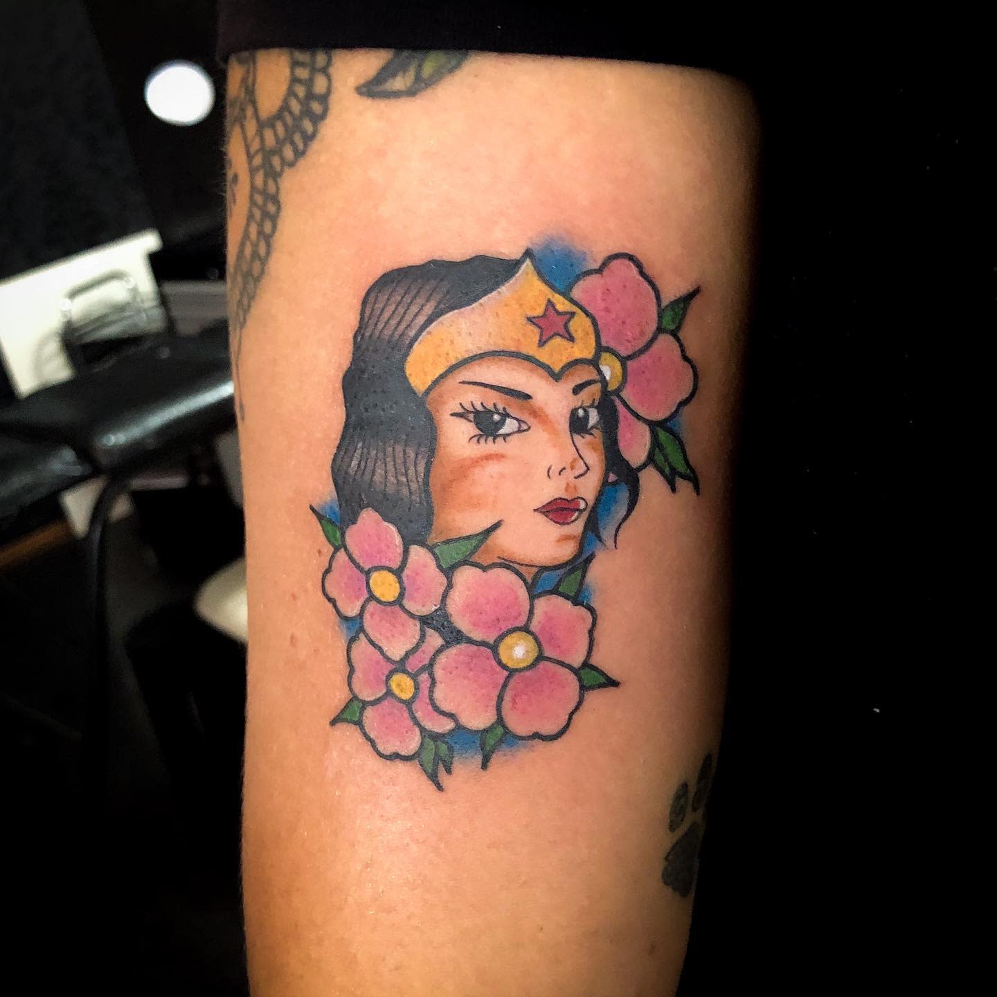 Traditional tattoo of Wonder Woman's face surrounded by pink flowers, blending classic comic style with floral elements