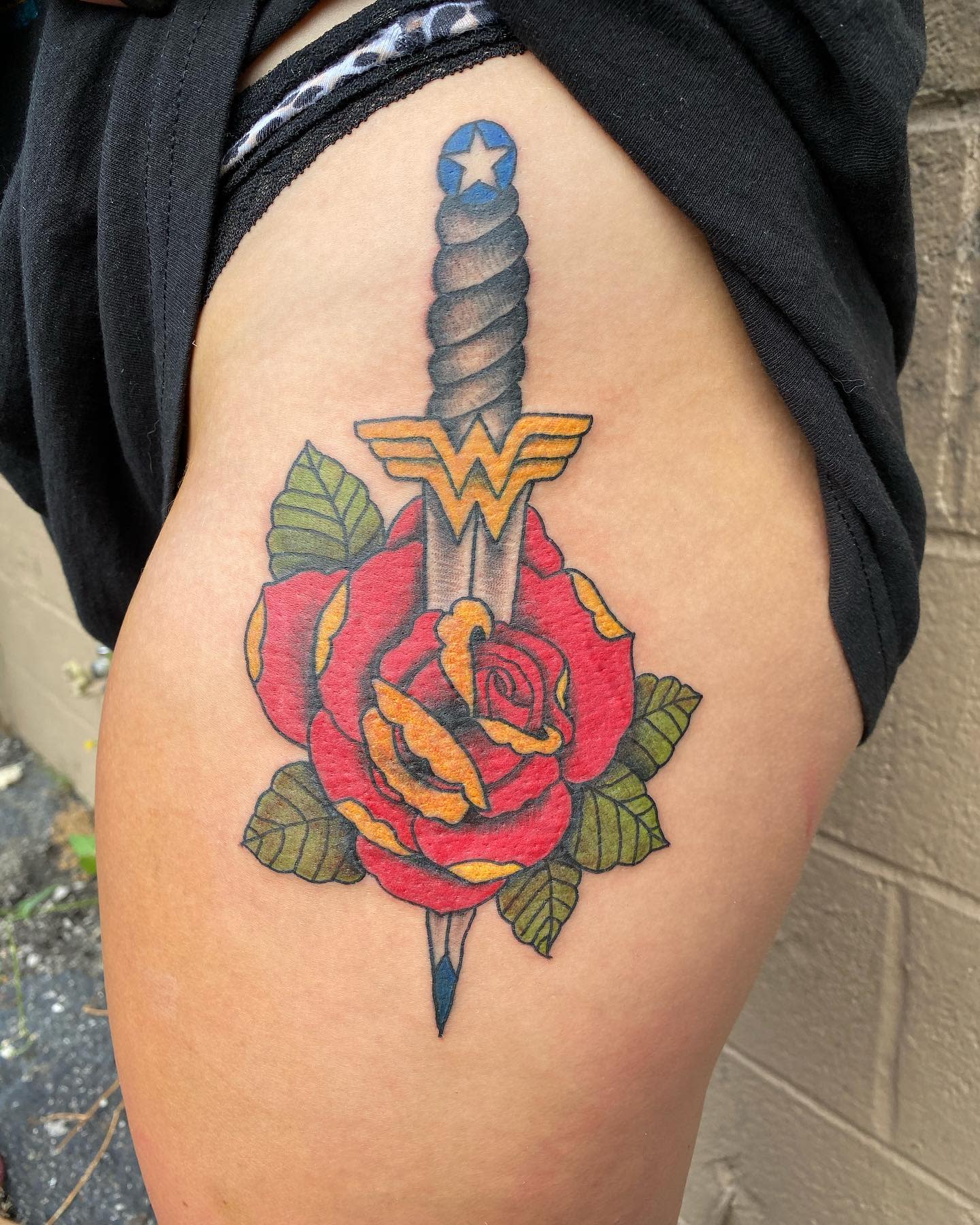 Traditional tattoo featuring Wonder Woman's sword piercing through a red rose, with her logo on the hilt