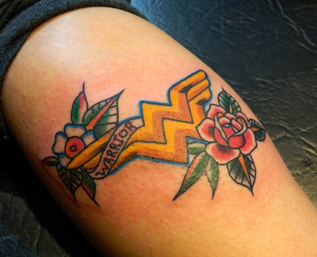 Traditional Wonder Woman tattoo featuring the 'WW' symbol surrounded by flowers and a banner that reads 'Warrior'