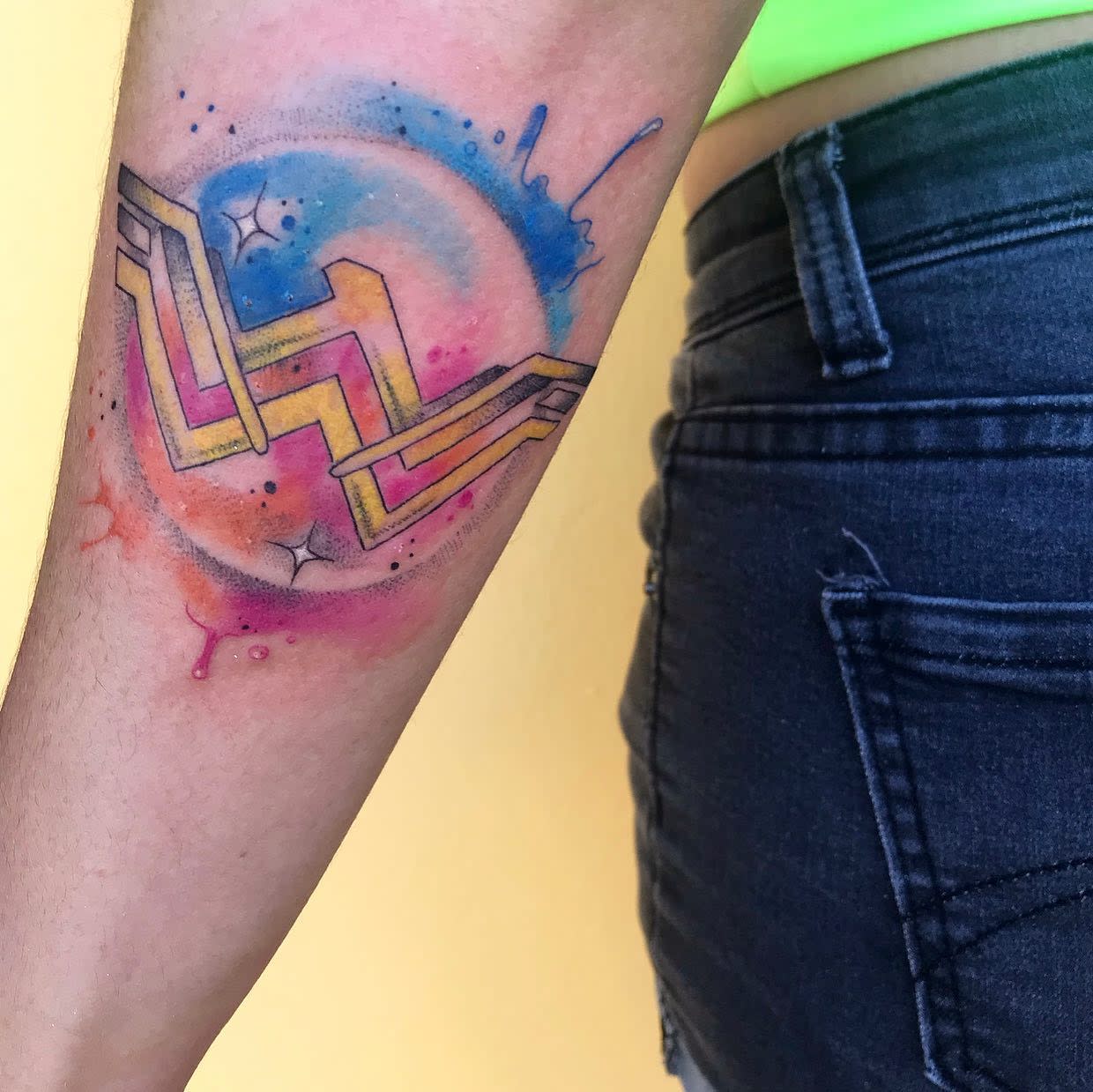 Wonder Woman logo tattoo with vibrant watercolor background, blending pinks, blues, and oranges for a dynamic look