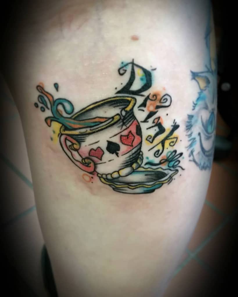 A playful tattoo of a teacup with a heart and spades, with letter 'Drink', liquid splashing from teacup on the saucer inspired by Alice in Wonderland