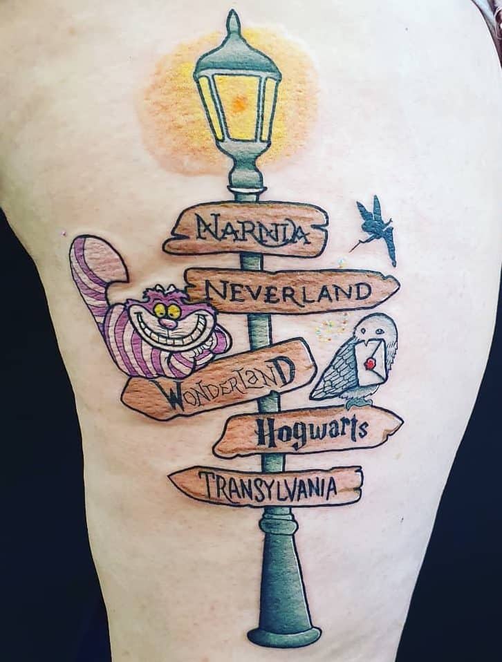 A whimsical tattoo featuring directional signs to fictional places like Narnia, Neverland, Wonderland, Hogwarts, and Transylvania, with the Cheshire Cat and owl holding a letter