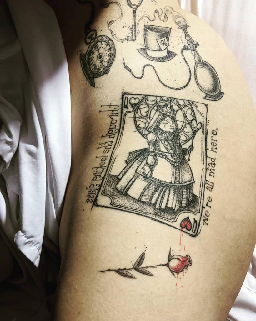 A detailed tattoo inspired by Alice in Wonderland, featuring the Mad Hatter's hat, pocket watch, hookah Queen of Hearts card, and the phrase "through the looking glass we're all mad here" with bleeding rose below