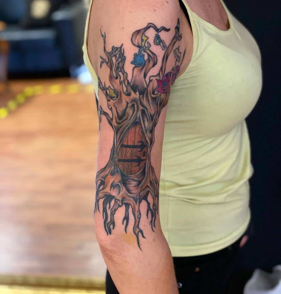 A creative tattoo of a tree with elements from Alice in Wonderland, including teacups, other things with tags, a Cheshire Cat, and a hidden door