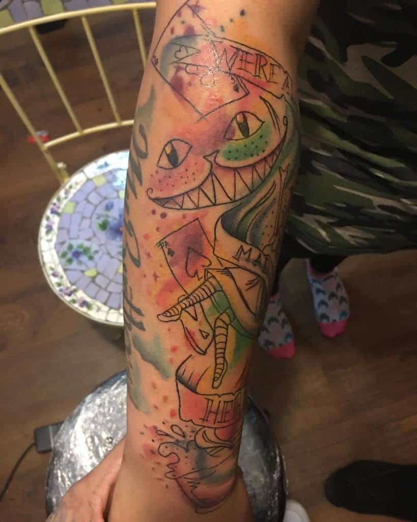 A vibrant tattoo of the Cheshire Cat from Alice in Wonderland, featuring bold colors, playing cards, and the phrase "We're All Mad Here"