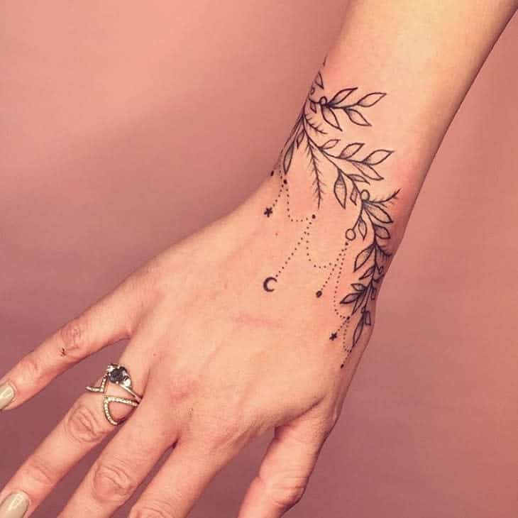 Delicate chandelier-style wrist tattoo featuring leafy vines, beaded chains, tiny stars, a crescent moon, and minimalist ornamental details