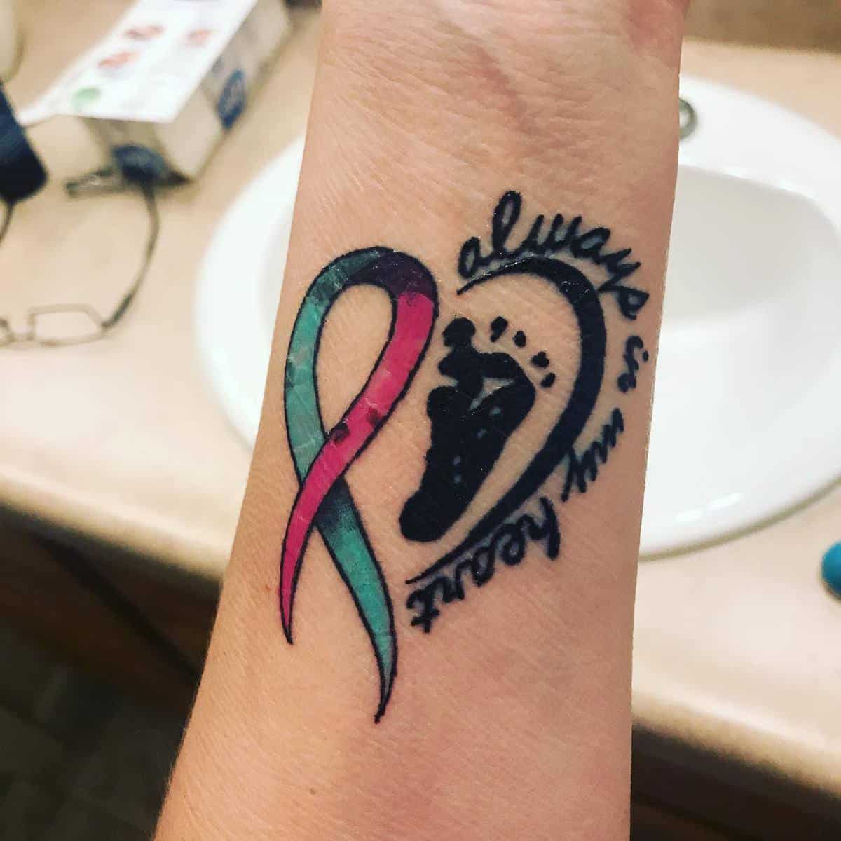 Tattoo of a teal and pink ribbon, a black baby footprint, and the words "always in my heart" on a wrist