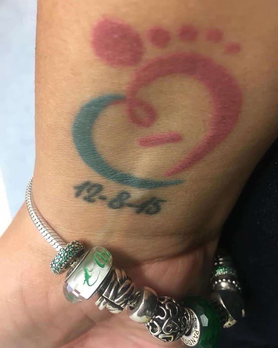 Tattoo of a red and blue heart with a footprint and date "10-8-15" on wrist
