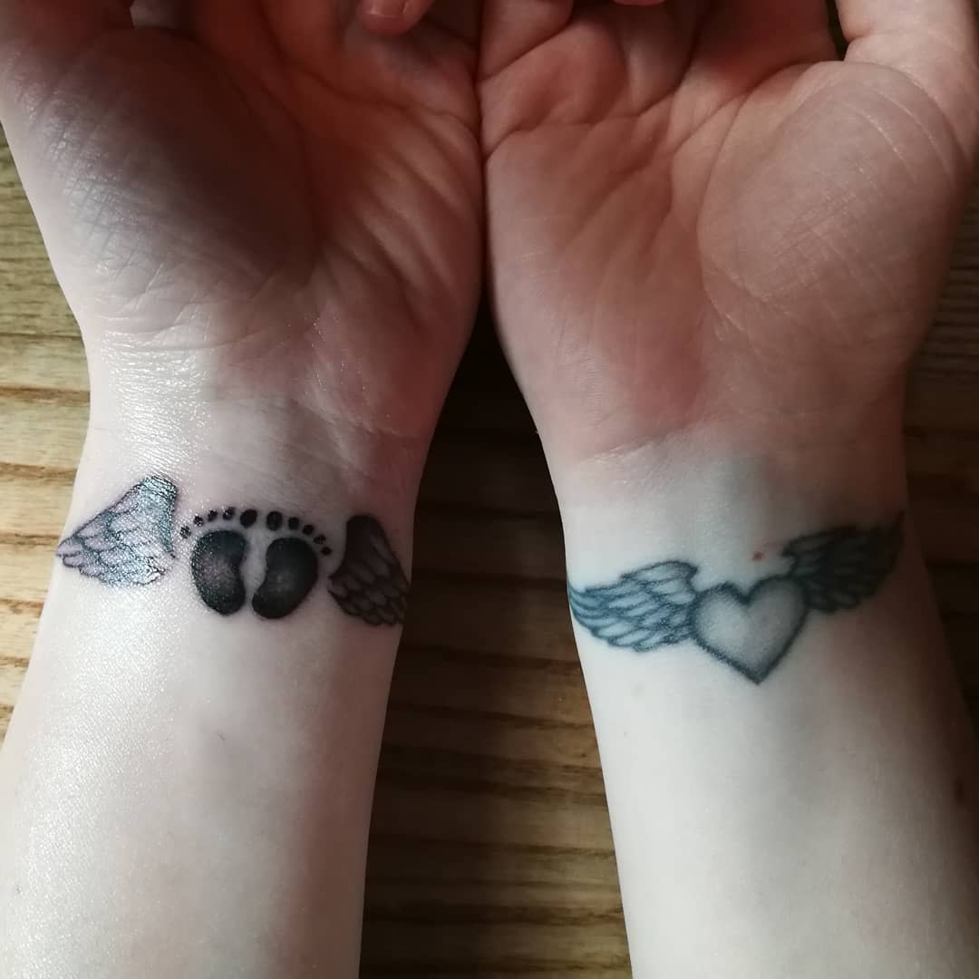 Two wrists with tattoos: left wrist with baby footprints and wings, right wrist with a winged heart