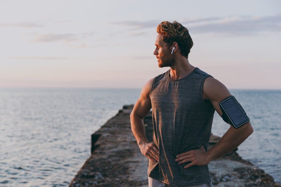 A man in workout gear stands on a pier at sunset, wearing earbuds—embracing mindful habits to restore balance and peace
