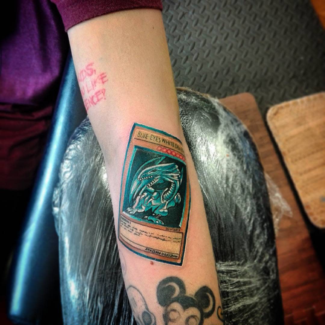 Tattoo of a Blue-Eyes White Dragon card on an arm with other tattoos visible