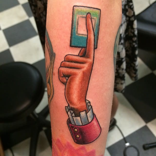 Tattoo of a hand pressing a green button on a finger on a forearm