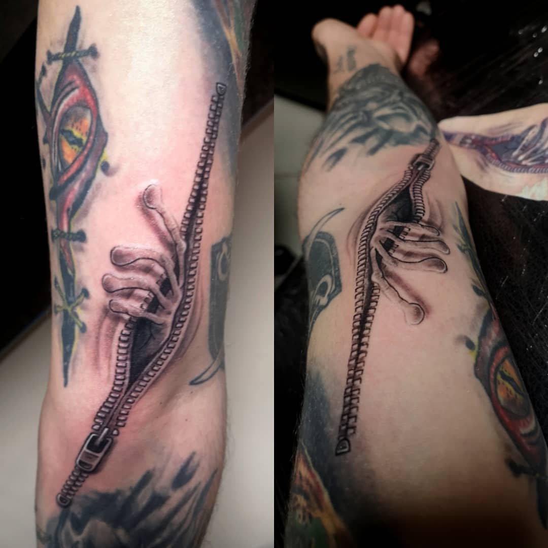 Tattoo of a realistic zipper with a skeletal hand emerging, inked on an arm