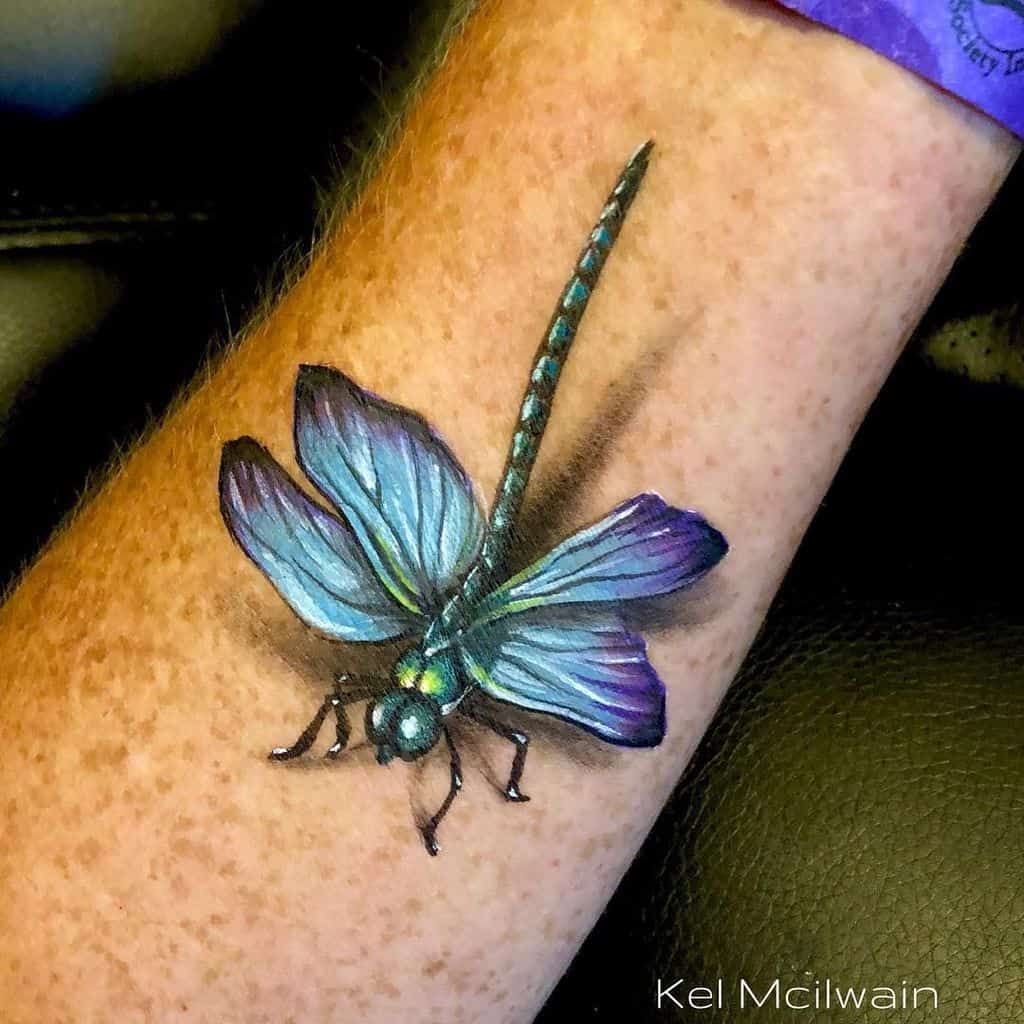 Hyper-realistic blue dragonfly tattoo with detailed wings and a shadow effect, placed on the forearm.